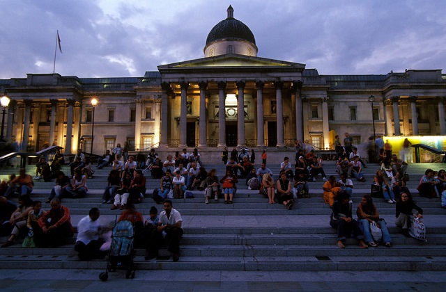 People sitting on steps in front of the National Gallery in Trafalgar Square at dusk.